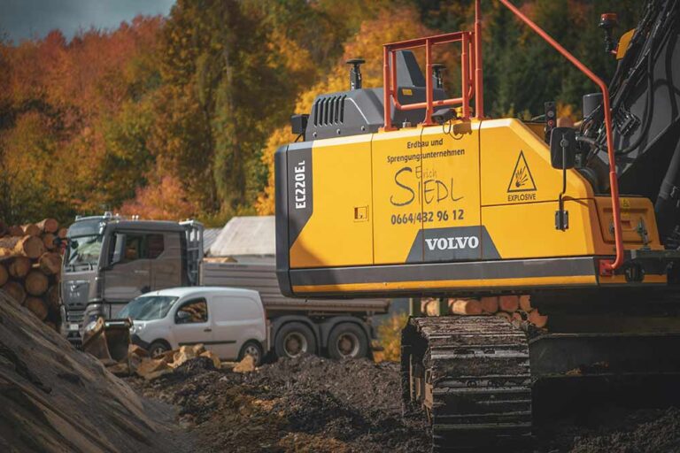 Teil des Baggers auf der Baustelle, im Hintergrund ein LKW und ein Auto.