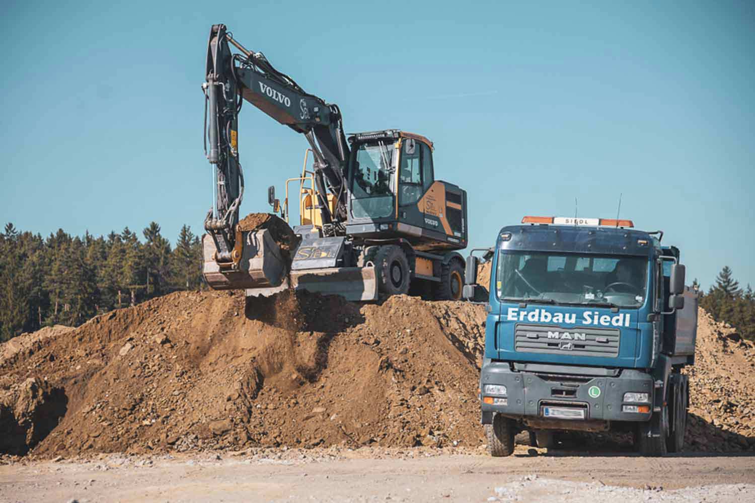Bagger von Erdbau Siedl auf einem Erdhaufen, daneben der blaue LKW.